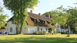 Een klassiek wit huis met rieten dak, omgeven door een verzorgd gazon en bomen.