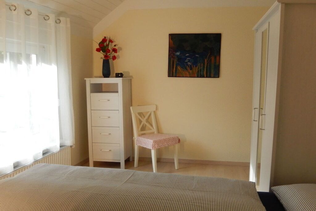 Un dormitori amb llit, cadira, prestatge i un quadre a la paret, i una finestra amb cortines.