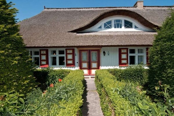 Ein traditionelles reetgedecktes Haus mit symmetrischer Fassade, einem zentralen Eingang mit roter Tür und einem Weg, der zum Eingang führt.