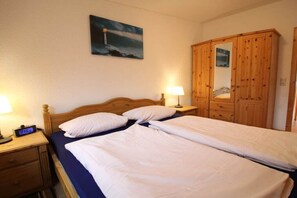 A double bed with white bedding, a wooden headboard, a nightstand with a lamp, a wooden wardrobe, and a painting on the wall. - 3-room apartment near the harbor - ground floor with terrace (Zingst)