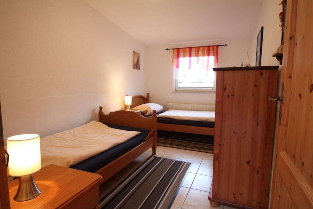 A room with two single beds, a wooden wardrobe, a bedside table with a lamp, and a window with a red and white curtain.
