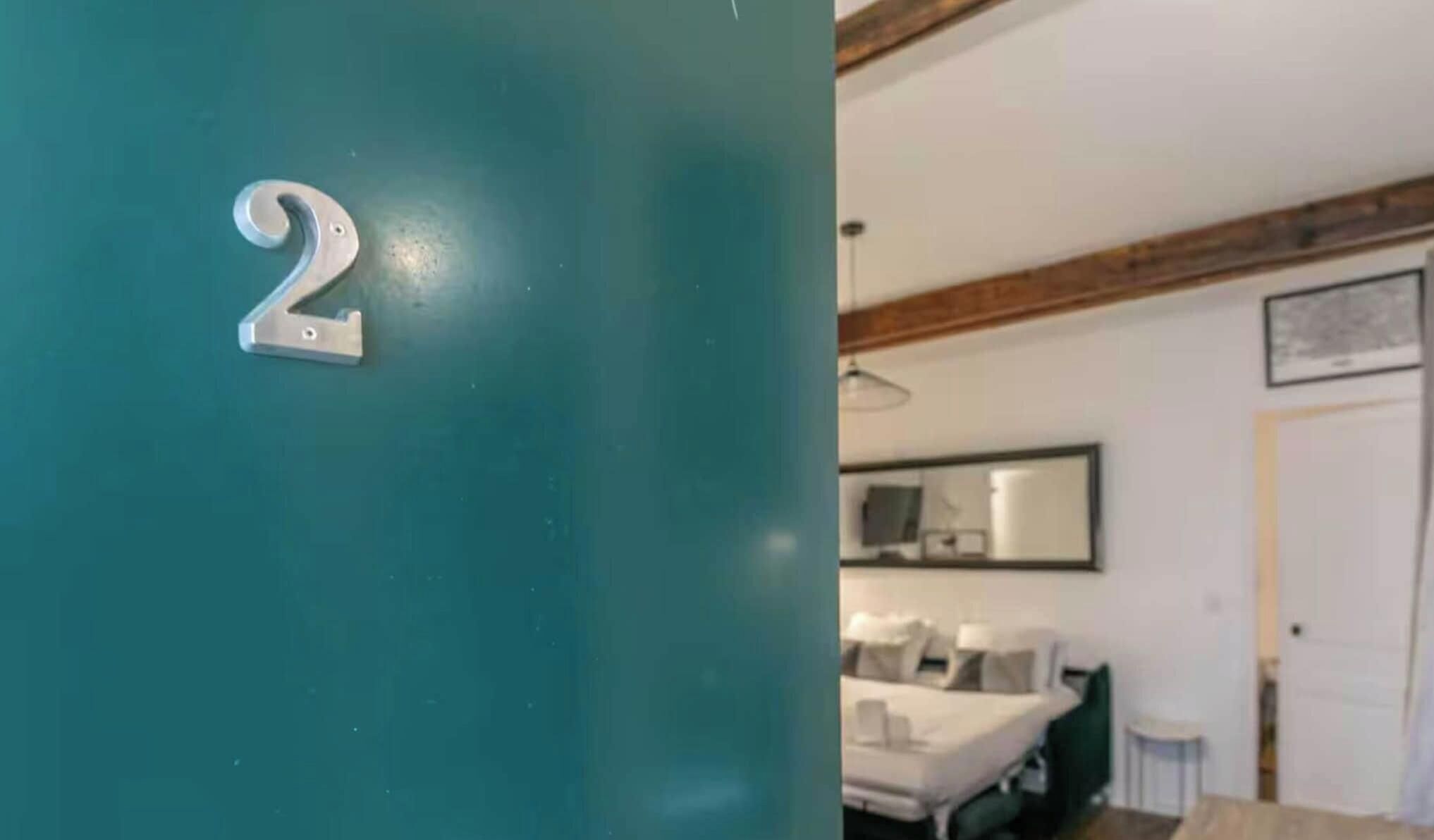 A room with a teal door marked with the number 2, a bed with white bedding, a bedside table, and a mirror on the wall.