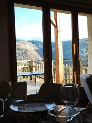 A dining area with a table and chairs, a window with a view of a mountainous landscape, and a wooden fence outside. - The Balcons du Haut-Doubs in the Heart of the Natural Park (Vaucluse)
