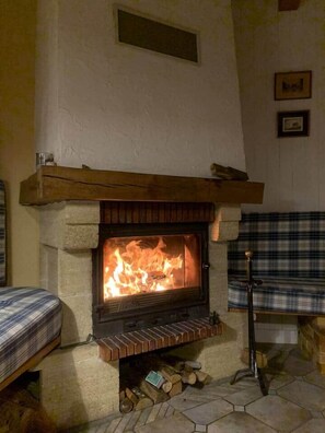 A stone fireplace with a fire, a wooden mantel, and a plaid bench. - The Balcons du Haut-Doubs in the Heart of the Natural Park (Vaucluse)