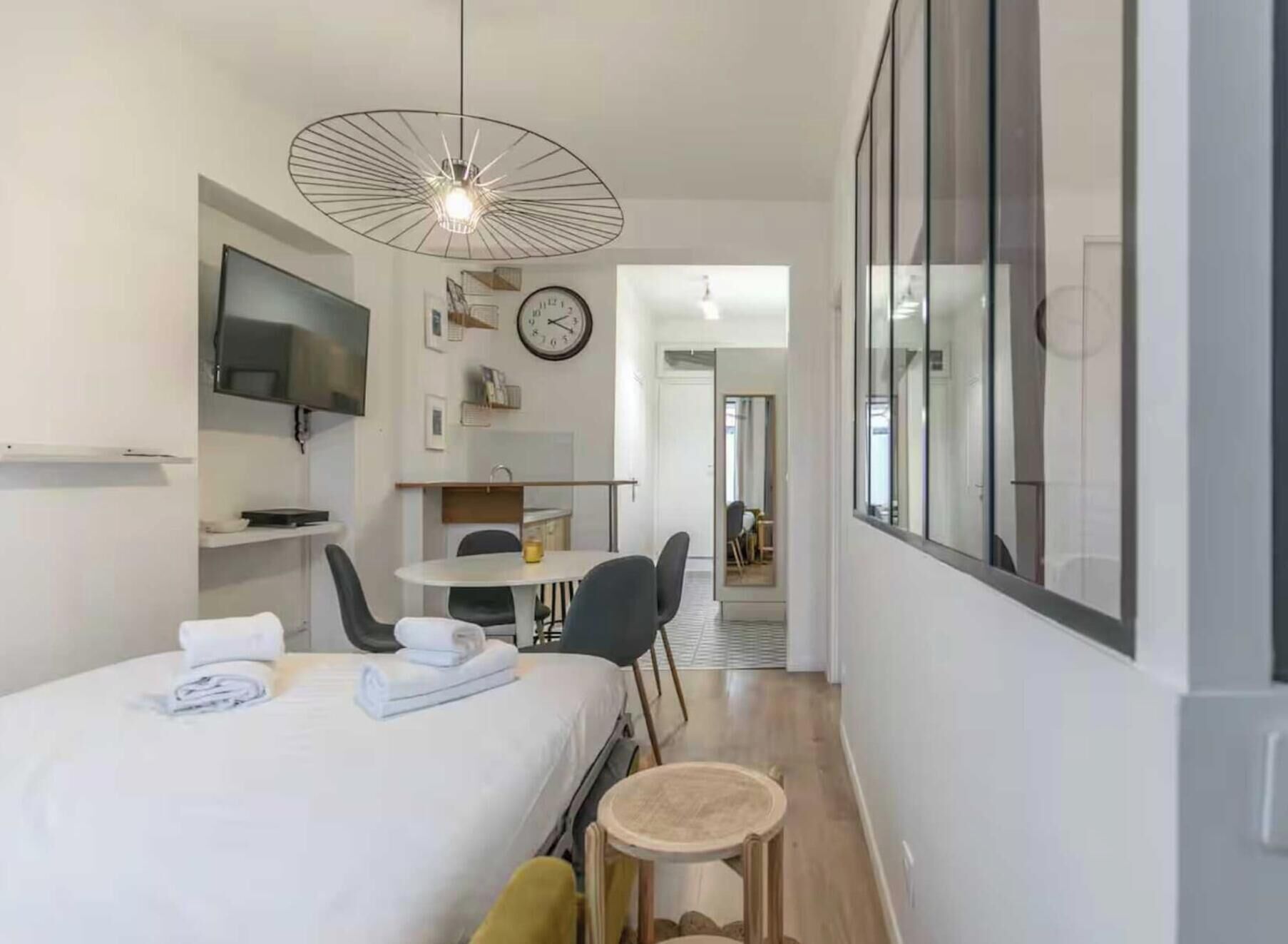 Quarto de hotel com cama, mesa pequena, cadeira, televisão, relógio e janela.