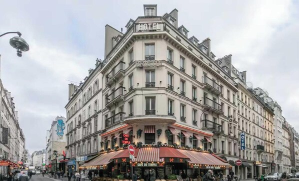 Um prédio de esquina com um letreiro 'HOTEL', um 'CAFE DU TEMPLE' com mesas ao ar livre e um letreiro 'BRIS'.