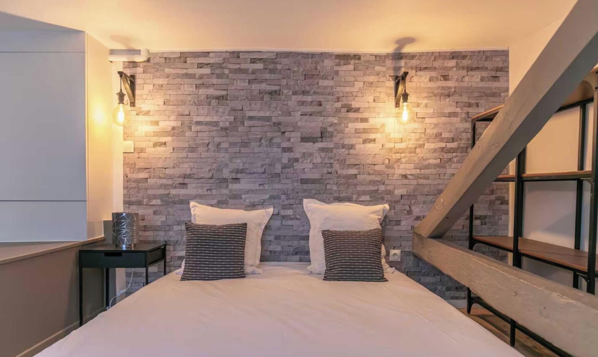 A bedroom with a brick-patterned wall, a bed with two pillows, a nightstand, and a staircase.