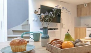 A kitchen area with a table displaying a muffin, a vase with dried flowers, a pear, and a fruit bowl containing an orange and kiwis. - Beautiful apartment- 1BR/4P - Oberkampf (Paris)