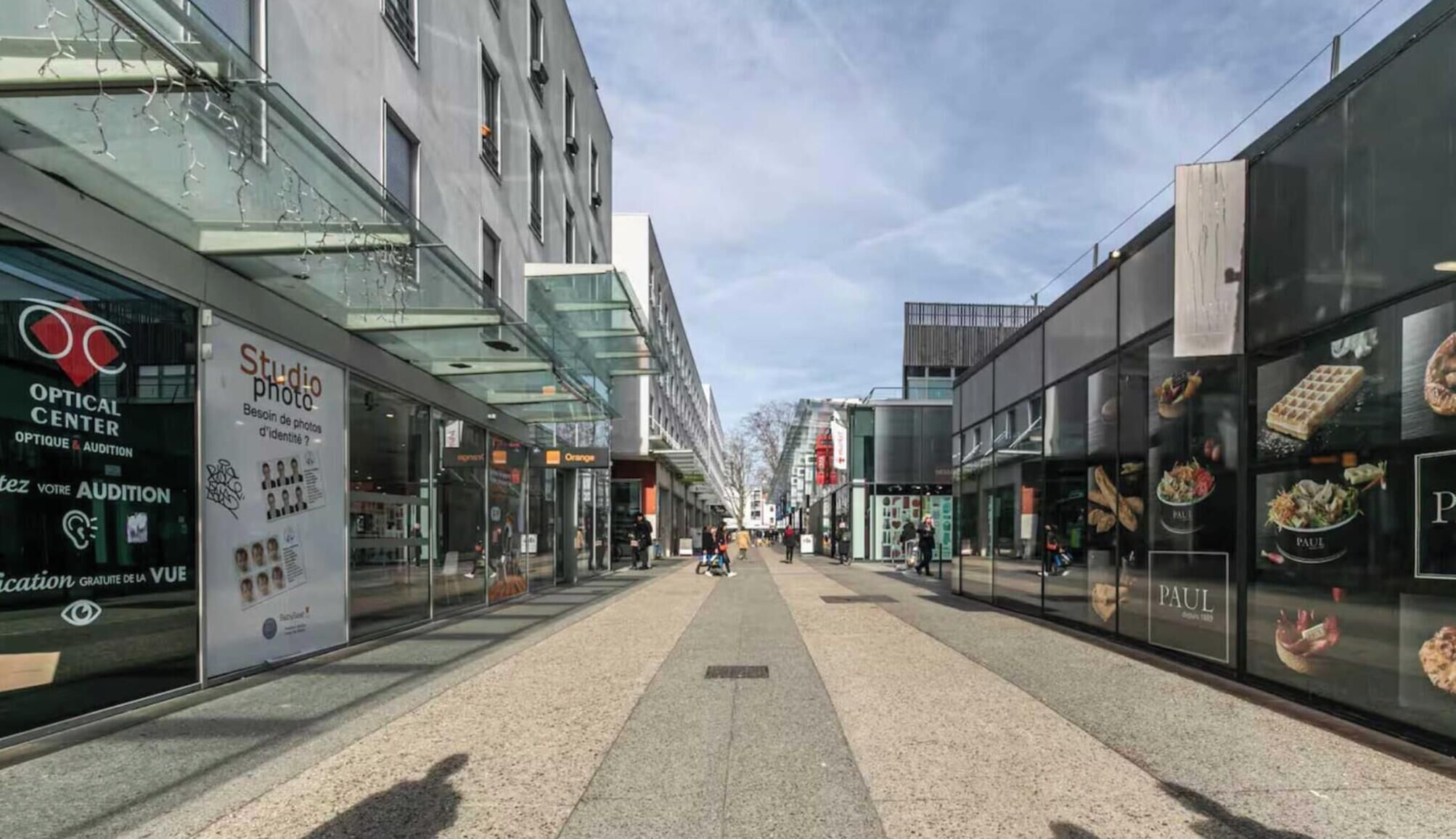 Una calle peatonal bordeada por edificios modernos, incluyendo una óptica y un restaurante.