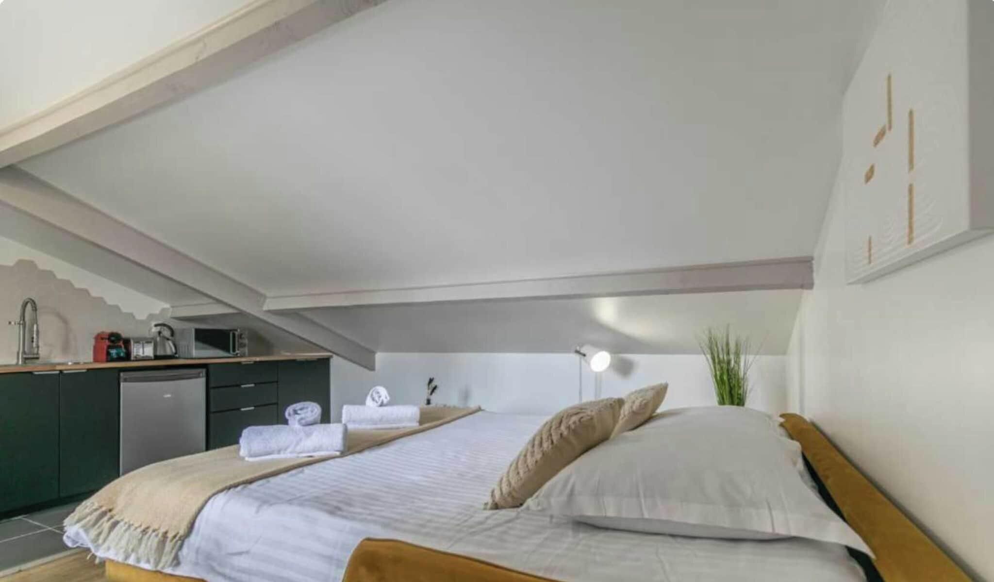 Quarto com cama, mesa de cabeceira, abajur e vista para uma área de cozinha.