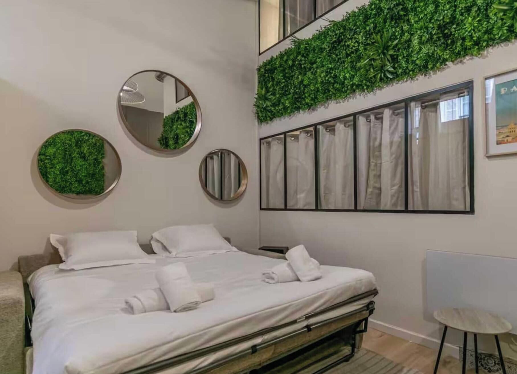 Une chambre d’hôtel avec un lit, deux miroirs ronds, un élément de décoration murale vert, une fenêtre avec des rideaux et une chaise.