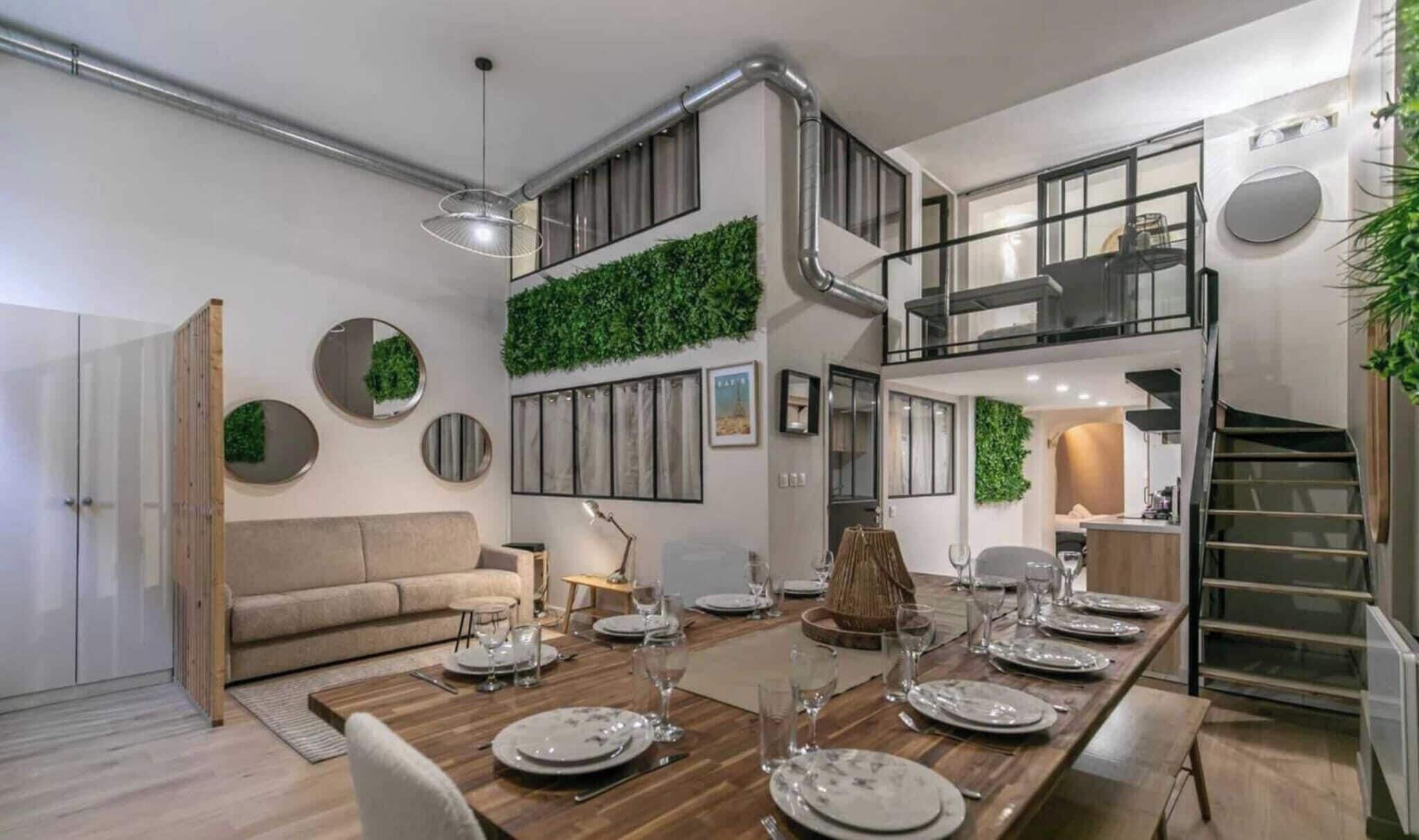 Un espace de repas moderne avec une longue table en bois dressée pour six personnes, un escalier et un coin salon agrémenté d’un canapé et de miroirs décoratifs.