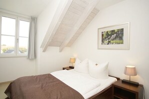 Een slaapkamer met een schuine kap, een bed met witte beddengoed, twee nachtkastjes met lampen en een ingelijste foto aan de muur.