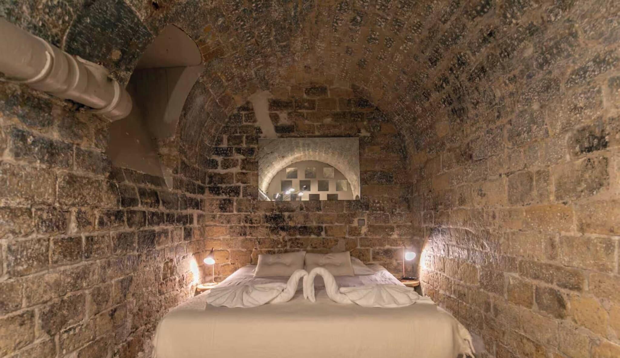 Una habitación de paredes de piedra con una cama, un pequeño ventanal de arco y una tubería a lo largo de la pared.