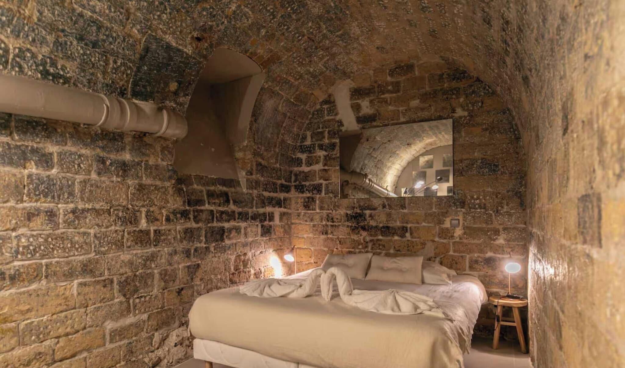 A stone-walled room with a single bed, a small table, and a mirror on the wall.