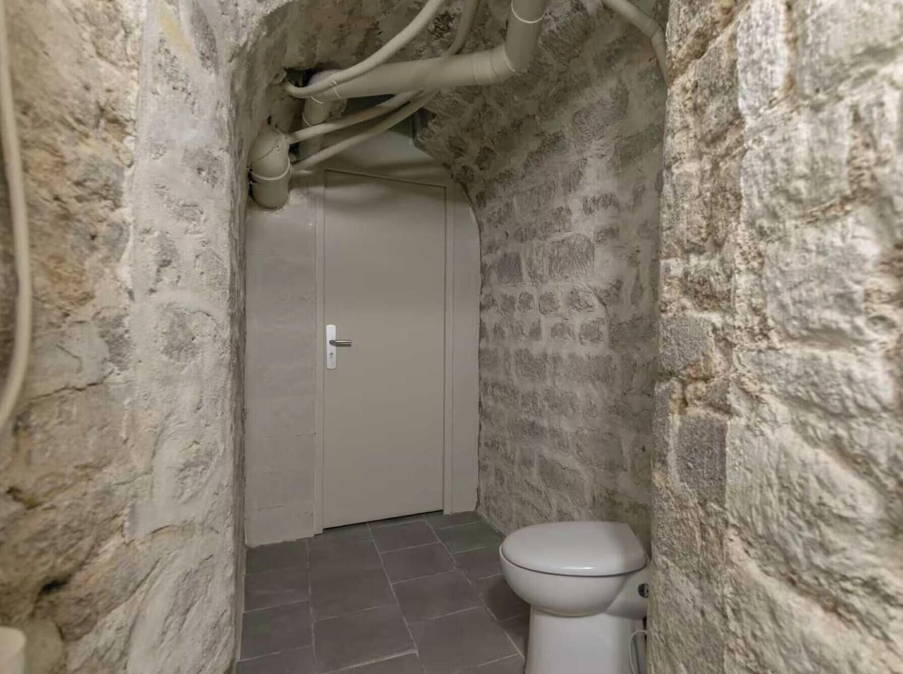 Een smalle badkamer met een wit toilet, betegelde vloer en stenen wanden.