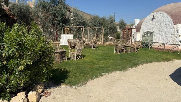 Un área de estar al aire libre con sillas y mesas de mimbre, rodeada de follaje y un edificio blanco con techo abovedado.