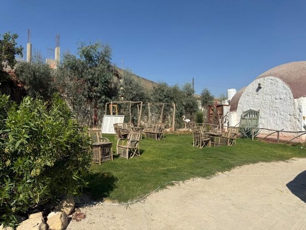 An outdoor seating area with wicker chairs and tables, surrounded by greenery and a white building with a dome-shaped roof.