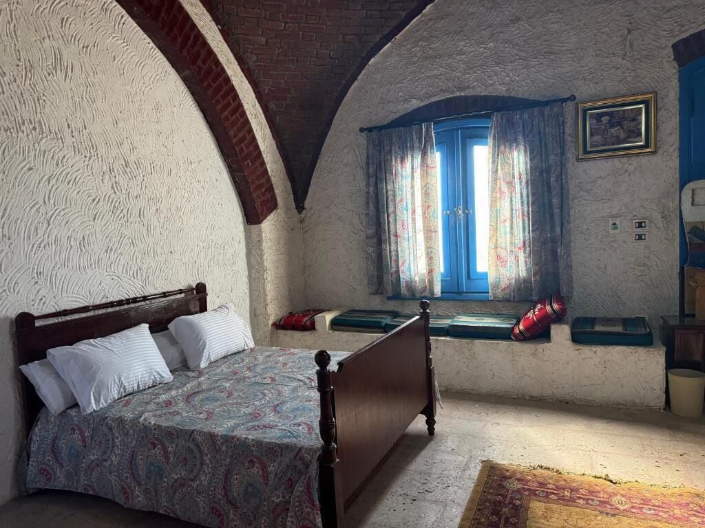A bedroom with a bed, a window with curtains, a wall clock, and a small table.