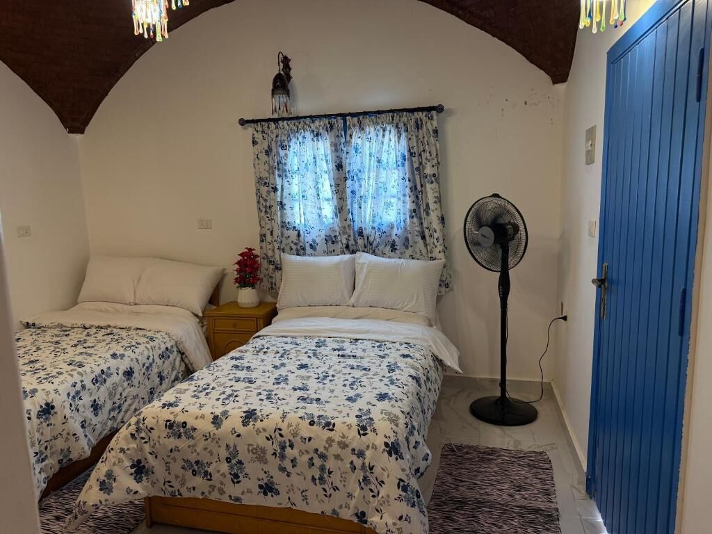 A bedroom with two beds, a standing fan, a blue door, and a window with curtains.