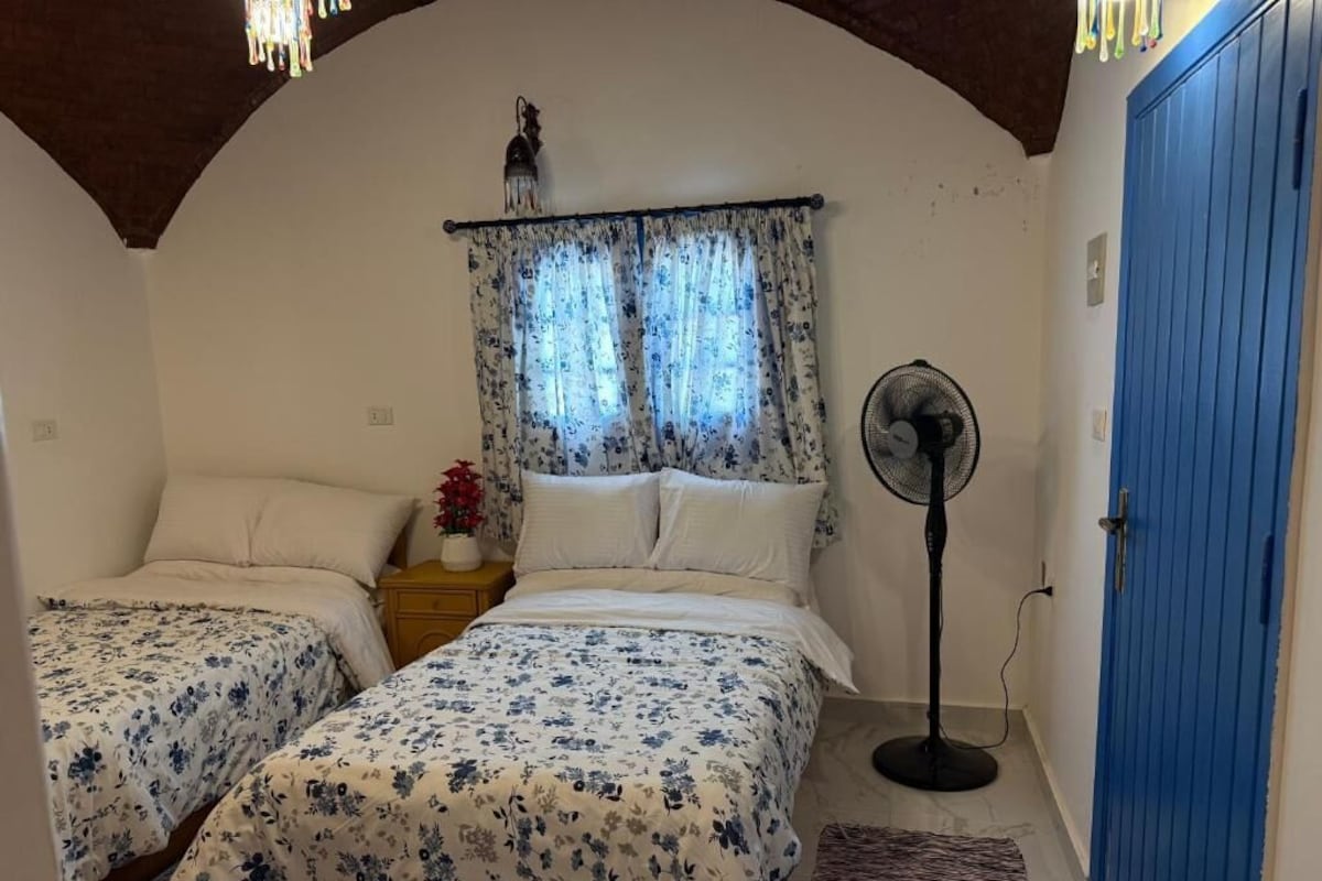 A bedroom with two beds, a standing fan, a blue door, and a window with curtains.