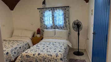 A bedroom with two beds, a standing fan, a blue door, and a window with curtains.