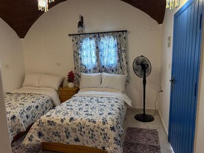 A bedroom with two beds, a standing fan, a blue door, and a window with curtains.