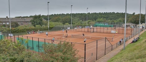 An outdoor tennis court with multiple players, surrounded by a fence and trees.