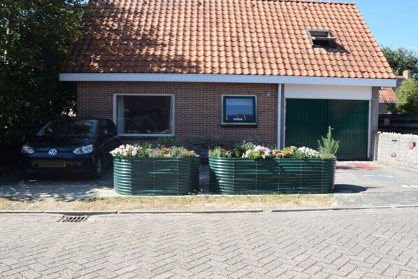 A small brick building with a tiled roof, a garage door, and two windows. There are two green planters with flowers in front of the building. - Holiday apartment with 2 bedrooms (Petten)