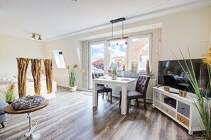 A modern living room with a dining area, a flat-screen TV, and a large window with a view. - Villa by the sea, home port (Sankt Peter-Ording)