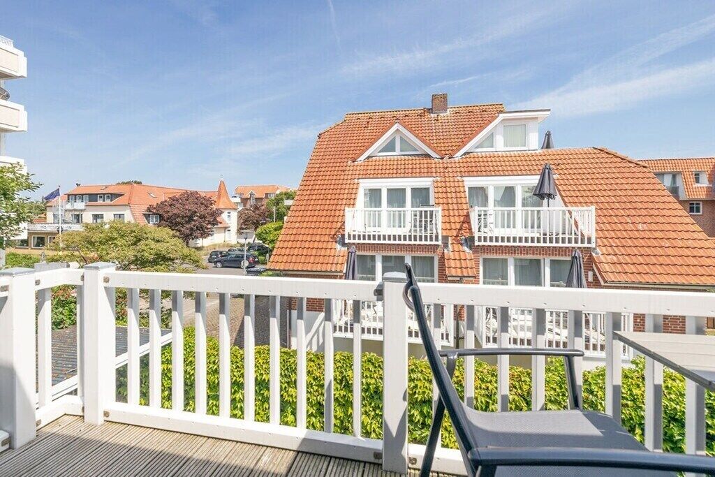 A balcony with a white railing, a lounge chair, and a view of a red-tiled building with balconies.