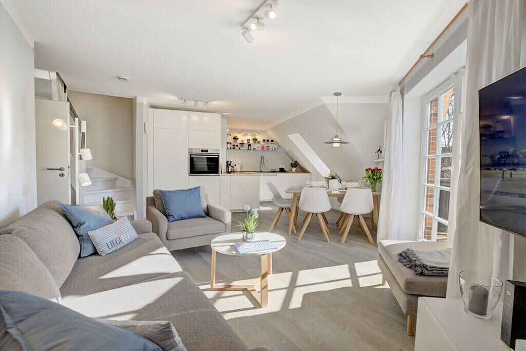 A modern living room with a sofa, coffee table, and TV. There is a kitchen area with white cabinets and a dining table.