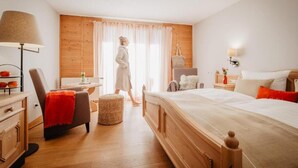 A bedroom with a wooden headboard, a bedside table with a lamp, a chair with a red throw, and a window with curtains. - Hotel Sankt Leonhard (Bad Birnbach)
