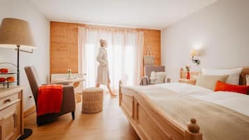 A bedroom with a wooden headboard, a bedside table with a lamp, a chair with a red throw, and a window with curtains.