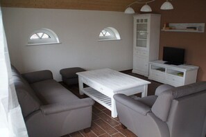A living room with a sofa set, a white coffee table, a cabinet, a television, and two arched windows. - Holiday home in the North Sea with 3 bedrooms (Nessmersiel)