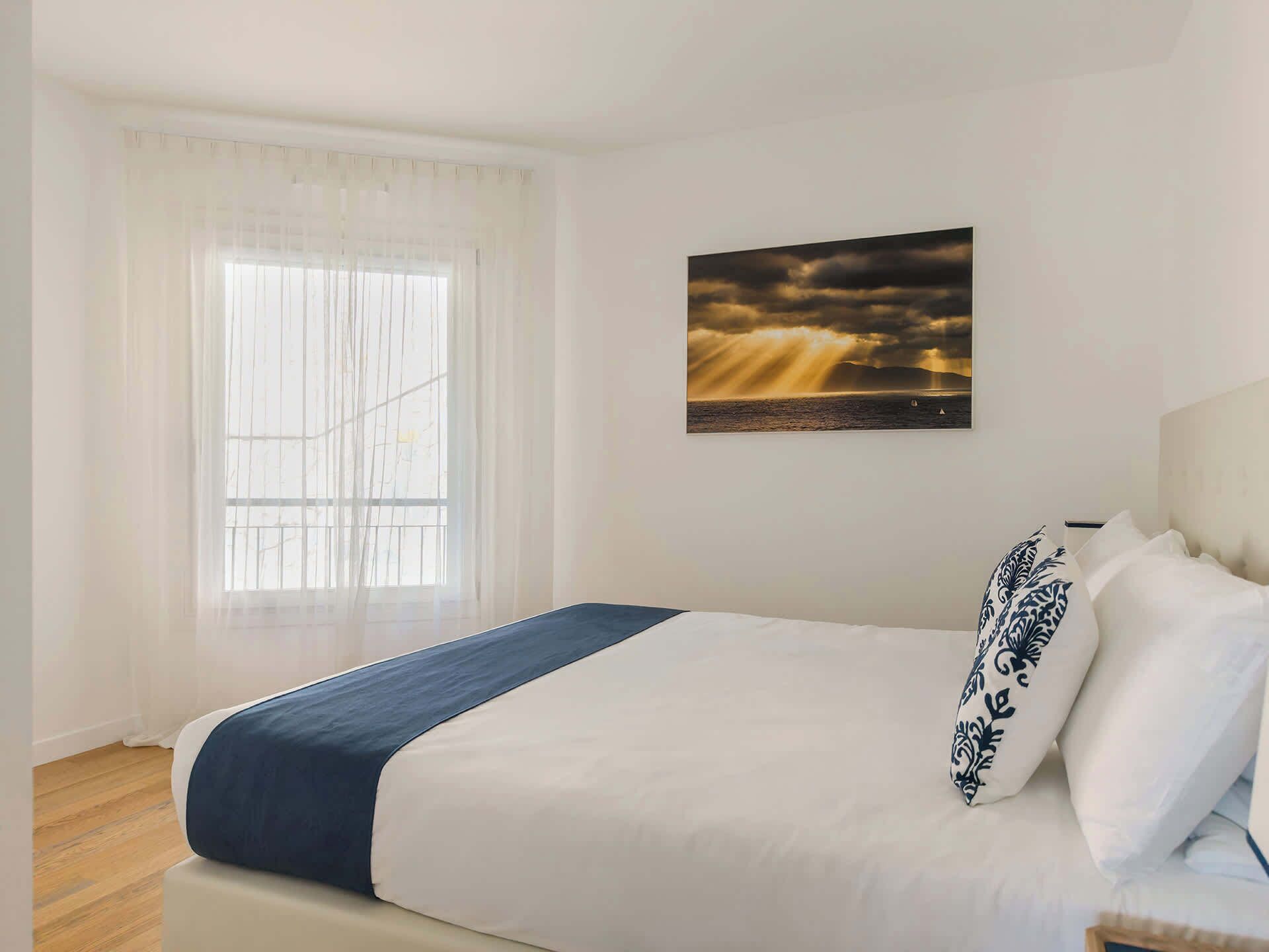 Une chambre à coucher avec un lit, une fenêtre ornée de rideaux légers et un tableau encadré accroché au mur.