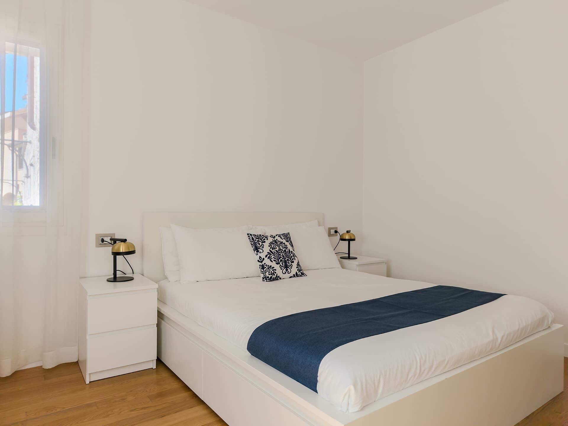 Ein Schlafzimmer mit einem Einzelbett, weißer Bettwäsche, einer blauen Tagesdecke, zwei Nachttischlampen und einem Fenster mit Vorhängen.