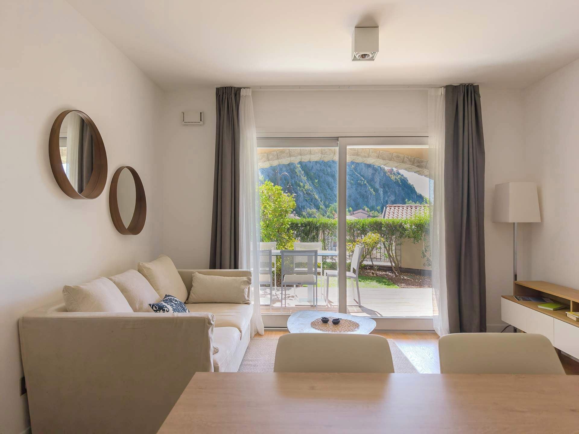 Un salon moderne avec un canapé, une table basse et une table à manger. Deux miroirs sont accrochés au mur, et une lampe est posée sur une table d’appoint. La pièce est dotée de grandes fenêtres avec des rideaux, offrant une vue sur une montagne et un jardin.