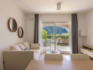 A modern living room with a sofa, a coffee table, and a dining table. There are two mirrors on the wall, and a lamp is on a side table. The room has large windows with curtains, offering a view of a mountain and a garden. - Giardino Segreto - Portopiccolo Apartments (Duino Aurisina)