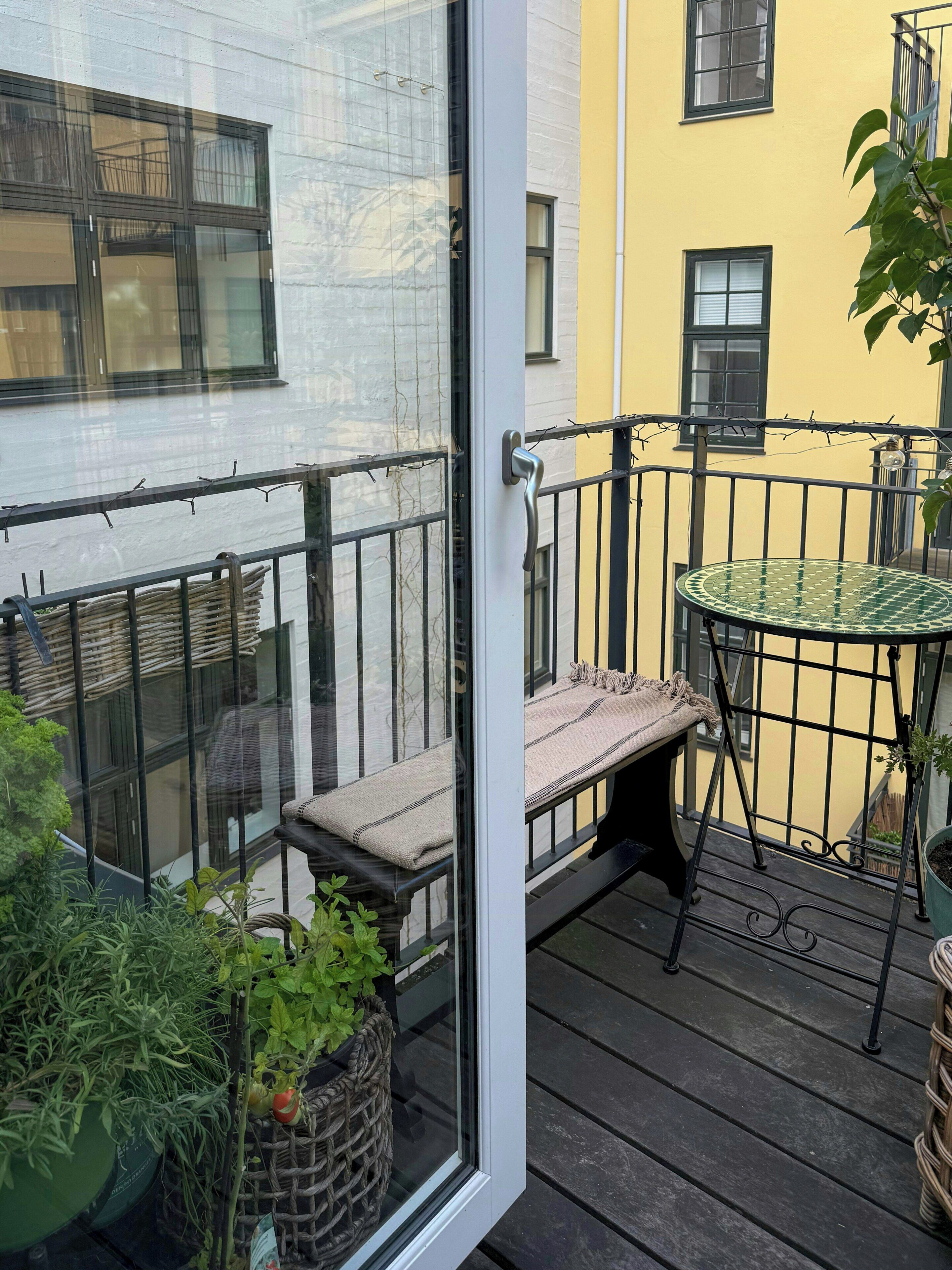 Ein Balkon mit Glastür, einem kleinen runden Tisch, einer Bank und Topfpflanzen.