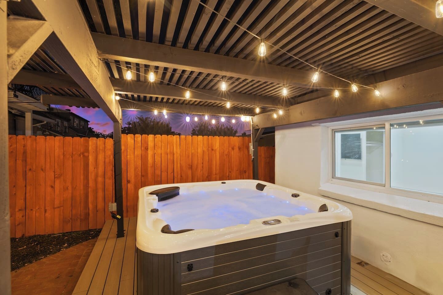 A hot tub under a wooden pergola with string lights, surrounded by a wooden fence and a window.