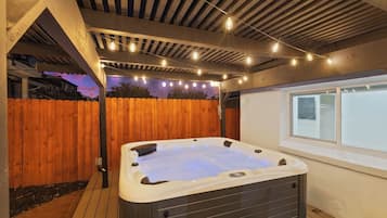 A hot tub under a wooden pergola with string lights, surrounded by a wooden fence and a window.