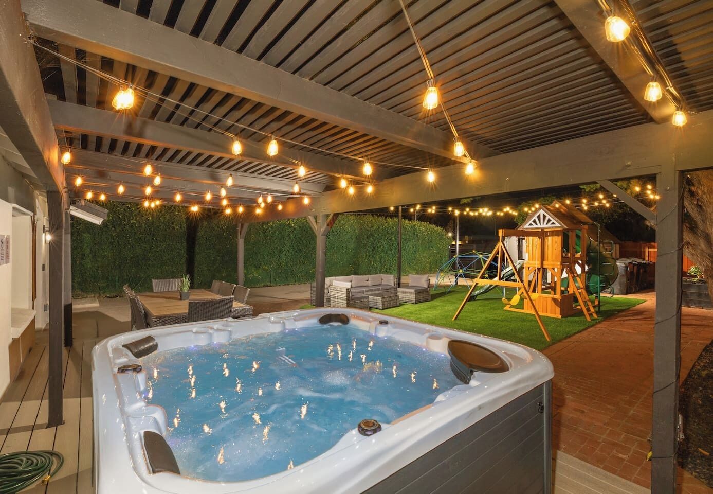 A hot tub under a covered patio with string lights, surrounded by outdoor furniture and a children's play structure.