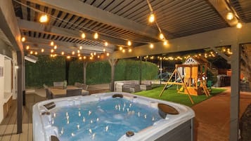 A hot tub under a covered patio with string lights, surrounded by outdoor furniture and a children's play structure.