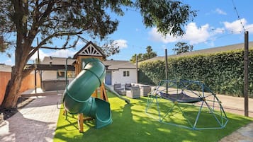 A playground with a slide and swings, surrounded by a lawn and a house.