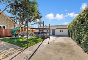 A residential backyard with a play area, including a slide and swings, a paved driveway, and a white house with a blue door.