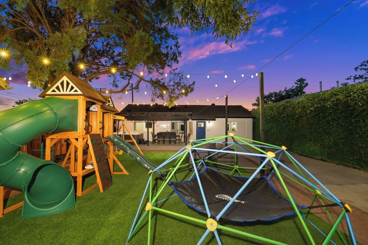 A playground with a slide and swings, a trampoline, and a house in the background.