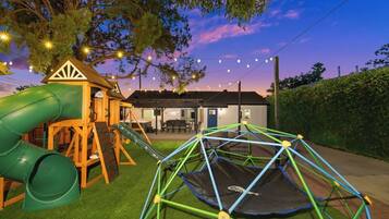 A playground with a slide and swings, a trampoline, and a house in the background.