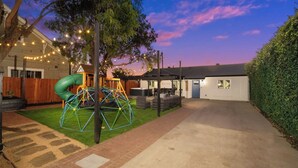 Un jardin arrière d’une maison, aménagé avec une aire de jeux, un espace de détente couvert et la maison elle-même.