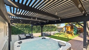 Un jacuzzi installé sous une pergola, avec un espace de jeux en arrière-plan.